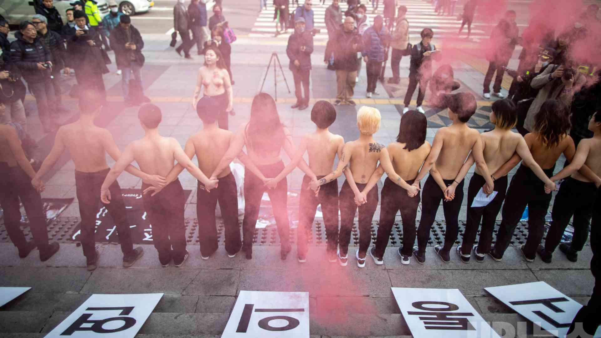 2020년 2월 14일 DxE가 진행한 시위 현장. 동물 강제 착유를 비판하며 서울 광화문 세종문화회관에 모였다. 시위 참여자들이 상의를 탈의하고, 등 뒤로 서로 손을 잡고 있는 동안 취재진과 행인이 사진을 찍고 있다. 현장 사진 제공 DxE.