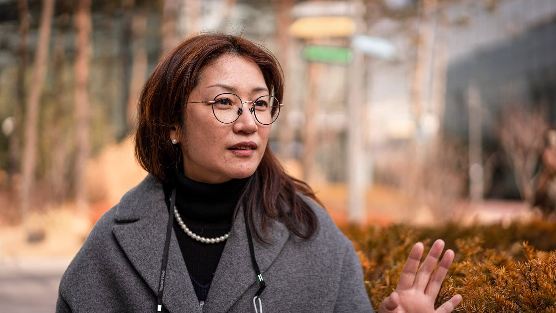 검정색 셔츠와 회색 코트를 입고 진주 목걸이를 한 지선씨가 왼손을 가슴 높이로 들고 있다.
